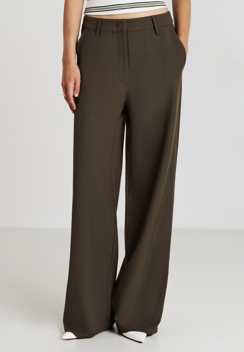 Pantalons larges en olive foncé, fabriqués à partir d'un tissu lisse. Présentent des poches avant, une taille ajustée et des chaussures pointues blanches.
