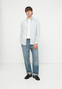 Νεαρός άνδρας στέκεται μπροστά σε λευκό τοίχο, φορώντας ανοιχτόγαλάζιο πουκάμισο πάνω από λευκή μπλούζα, φαρδιά μπλε τζιν με γυρισμένα μπατζάκια και μαύρα μοκασίνια.