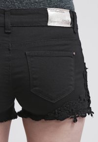 Černé džíny šortky s třepeným lemem, ozdobnými krajkovými detaily po stranách a stříbrným logem značky na pásku.