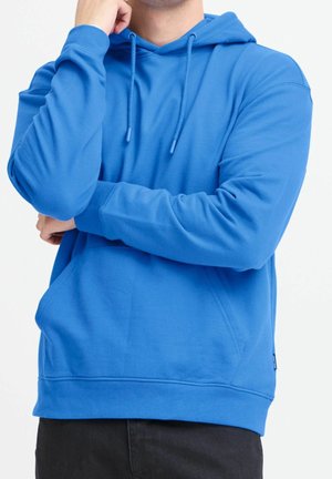 Homme portant un sweat à capuche bleu vif avec poche avant et cordons, bras croisés avec une main touchant le menton, sur fond blanc uni.
