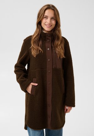 Femme aux longs cheveux bruns portant un manteau en polaire marron foncé boutonné sur un jean bleu, souriante et posant avec une main dans la poche.