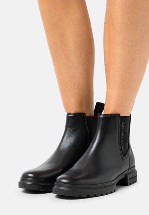 Bottes cheville en cuir noir avec panneaux latéraux élastiques, bout rond, semelle texturée, et détail logo à l'arrière du talon et sur l'élastique.