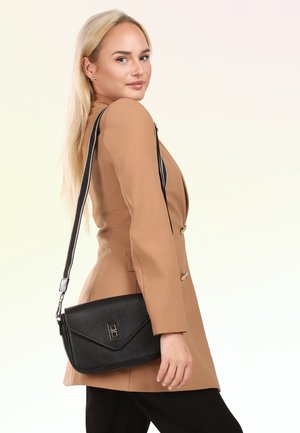 Blonde vrouw in camelkleurige blazer en zwarte broek draagt een zwarte leren crossbodytas met zilveren gesp, kijkt over haar schouder.