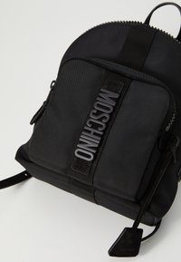 Čierny batoh Moschino s prednou zipsovou kapsou a názvom značky v vystúpených písmenách na látkovej páske, umiestnený na jednoduchom svetlom pozadí.