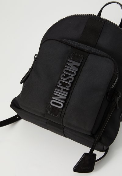 Μαυρη τσάντα πλάτης Moschino με μπροστινή τσέπη με φερμουάρ και την επωνυμία σε ανάγλυφα γράμματα πάνω σε μια υφασμάτινη λωρίδα, σε έναν απλό φωτεινό φόντο.