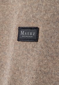 Beige stickad tyg med en svart rektangulär sydd etikett som visar "MAERZ MÜNCHEN" i vita versaler.