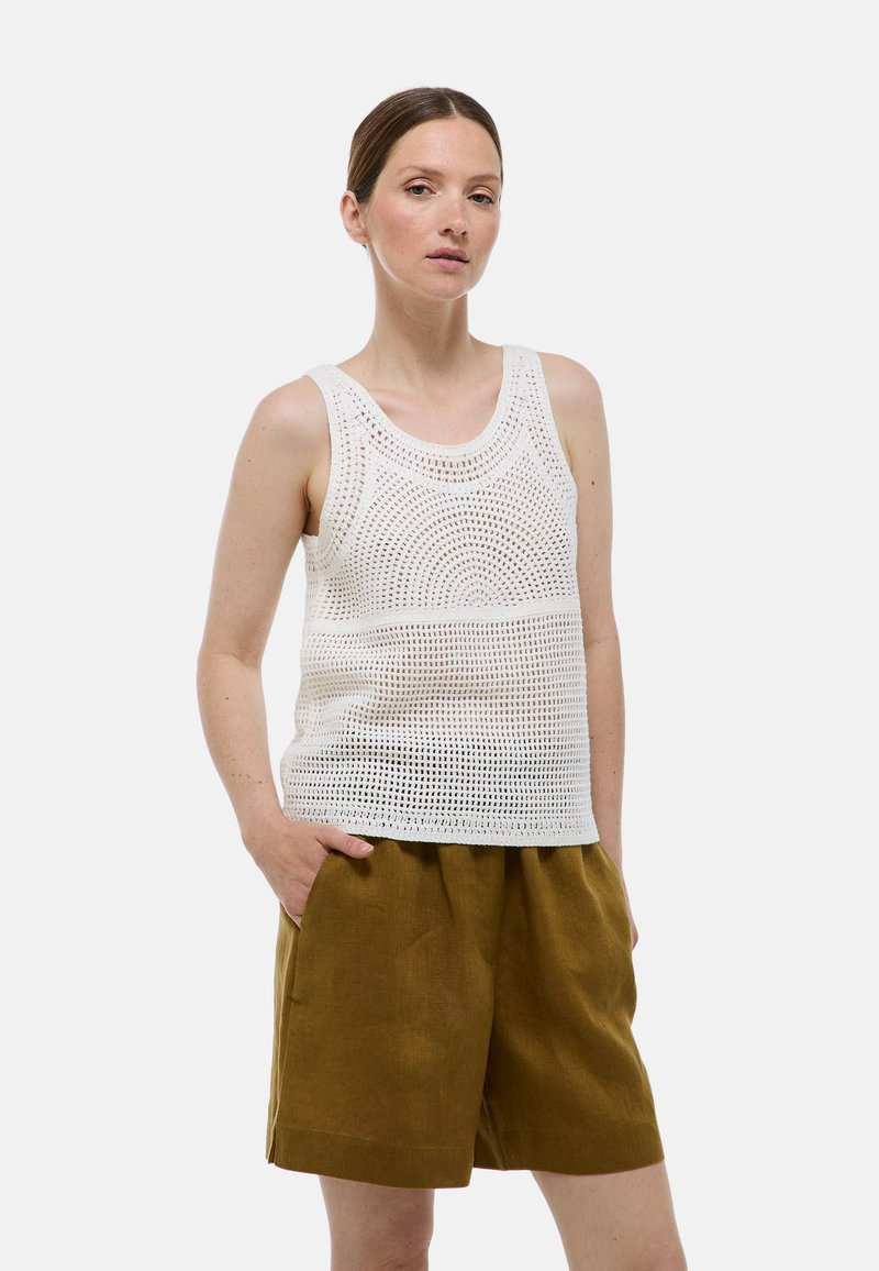 Débardeur en crochet blanc à encolure ronde, avec un design en tricot ajouré. Assorti à un short en lin marron, les mains dans les poches.