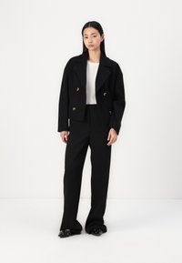 Black double-breasted jacket with gold buttons, paired with a textured white top and high-waisted black trousers. Flat black shoes with accents.