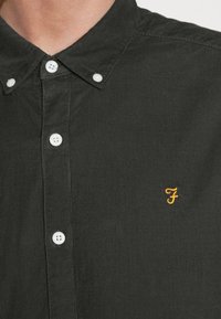 Czarna koszula sztruksowa z kołnierzykiem na guziki, z białymi guzikami i małym żółtym haftowanym logo po lewej stronie.