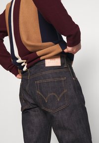 Tummansiniset farkut, joissa on kontrastiväriset keltaiset ompeleet, ja vaaleanpunainen nahkainen etiketti, jossa lukee "EDWIN". Yllä on värikkäästi raidoitettu neulepaita.