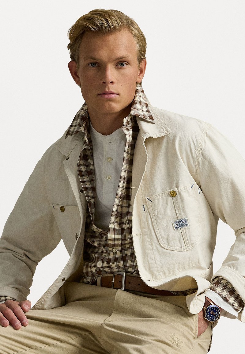 Chaqueta color crema con costuras azules y un bolsillo lateral sobre una camisa a cuadros marrón y blanca. Pantalones de color claro y un reloj azul.