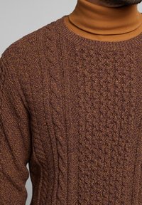 Brauner Strickpullover mit strukturierten Mustern, einschließlich Zopf- und Fischgrätmuster, kombiniert mit einem braunen Rollkragenpullover darunter.