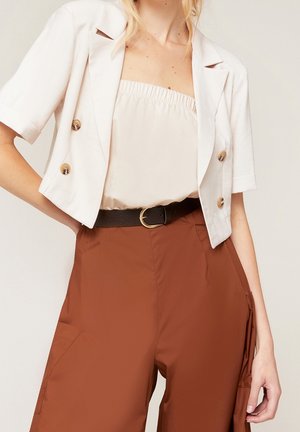 Kvinde iført en hvid cropped blazer, beige stropfri top, brune bukser med høj talje med sidelomme og et sort bælte med guldspspænde.