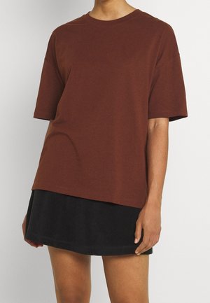 Persoon die een losse bruine korte mouwen shirt draagt over een korte zwarte rok, staand tegen een effen lichte achtergrond.
