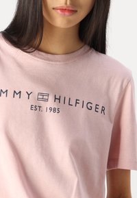 Lyserød bomuld t-shirt med korte ærmer, prydet med et mørkeblåt logo, der står "TOMMY HILFIGER EST. 1985" på tværs af brystet.