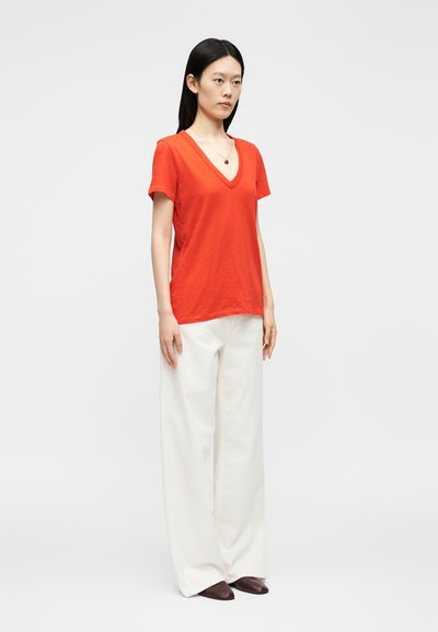 Γυναίκα στέκεται φορώντας πορτοκαλί κοντομάνικη μπλούζα με V λαιμόκοψη, λευκό παντελόνι με φαρδιά μπατζάκια, καφέ παπούτσια και κολιέ με μενταγιόν, μπροστά σε απλό φόντο.