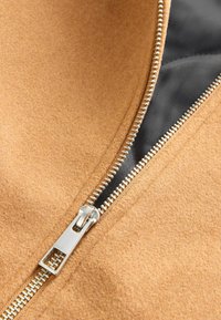 Tejido de lana color beige con un zipper metálico y forro negro, que presenta una textura suave y costuras visibles a lo largo de la unión.