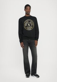 Črna majica s kapuco z velikim zlatim logotipom Versace Jeans Couture. Ustvarjena v kombinaciji s temno sivimi kavbojkami in črnimi športnimi copati. Preprost, sproščen dizajn.