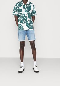 Camicia a maniche corte con motivi di foglie verdi su tessuto bianco, abbinata a pantaloni corti in denim blu chiaro arrotolati e scarpe bianche con dettagli neri.