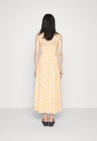 Um vestido maxi com estampado floral amarelo, com alças espaguete ajustáveis, corpete ajustado e saia fluida. Tecido texturizado com padrões florais visíveis.