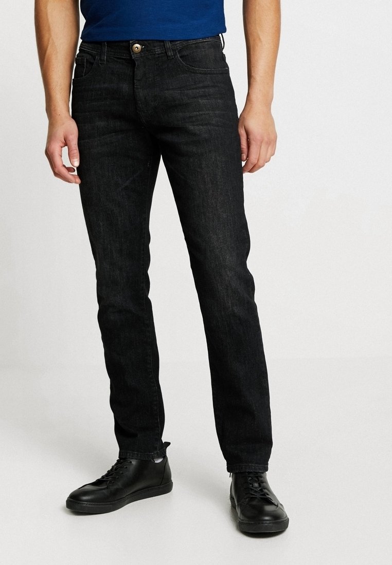 Personne portant un jean noir slim, des baskets noires et une chemise bleue, debout devant un fond clair uni.