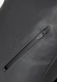 Borsa in pelle nera con una texture liscia, un logo impresso e una cerniera con linguetta, arricchita da sottili dettagli di cucitura.