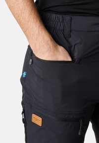 Black hiking pants made of durable fabric, featuring a zippered pocket, an elastic waistband, and a Swedish flag accent near the pocket.