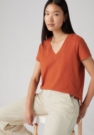 Rood-oranje v-hals t-shirt in een zachte stof, korte mouwen, relaxte pasvorm, met een klein stoffen label aan de onderzijde, gecombineerd met crèmekleurige broek.