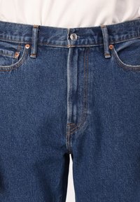 Temno modri jeans iz denima imajo klasičen dizajn s petimi žepi, bakrene dodatke in vidno oranžno šivanje za kontrast.