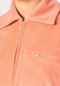 Veste zippée couleur corail en tissu éponge doux, dotée d'un col et d'une petite poche zippée sur la poitrine gauche.