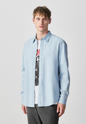 Junger Mann, der ein hellblaues Hemd über einem weißen T-Shirt mit Grafik trägt, stehend vor einem schlichten grauen Hintergrund.