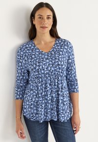 Blauwe bloemen top met driekwartmouwen, V-hals, geplooide taille en variërende tinten blauw en zwart in patronen. Draag met blauwe jeans.