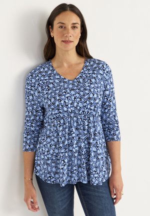 Bluse mit Blumenmuster in Blau, mit drei Viertelärmeln, V-Ausschnitt, geraffter Taille und unterschiedlichen Schattierungen von Blau und Schwarz. Getragen mit blauen Jeans.