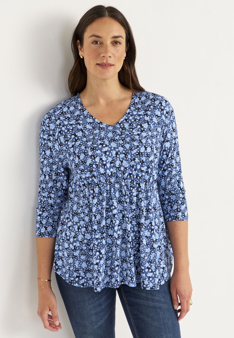 Blå blomstret top med trekvartærmer, V-hals, samlet talje og varierede nuancer af blå og sort mønstre. Båret med blå jeans.