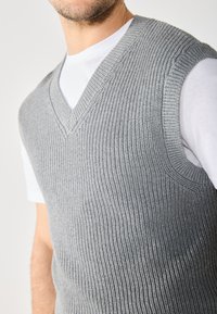 Grå strikket ermeløs vest med V-halsdesign, med teksturert ribbing og båret over en hvit T-skjorte.
