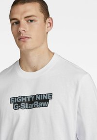 Hvit bomulls-T-skjorte med en fremtredende svart og grå grafisk tekstlogo som leser "EIGHTY NINE G-StarRaw" på forsiden.