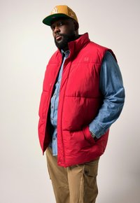Rote Steppweste über einem hellblauen Jeanshemd, kombiniert mit Khakihosen und einer gelben Baseballmütze. Die Weste hat einen Reißverschluss vorne und Taschen.