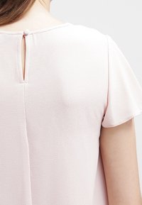 Ljusrosa kortärmad topp med texturerad tyg, nyckelhål tillbaka detalj med knappstängning och mjuk, flödande silhuett.