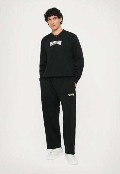 Sweatshirt noir et jogging assorti en tissu doux ; présente un logo blanc sur la poitrine et sur la jambe ; le mannequin porte des baskets blanches.