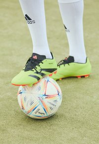Bota de fútbol verde y negra con acentos naranjas, que presenta una superficie texturizada, cordones y un logotipo, descansando sobre un balón de fútbol con un diseño estampado.