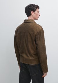 Brun ruskindjakke med klassisk krave, lige snit og rygsømsdetaljer. Bæres med mørke jeans, hvilket fremhæver en glat tekstur.