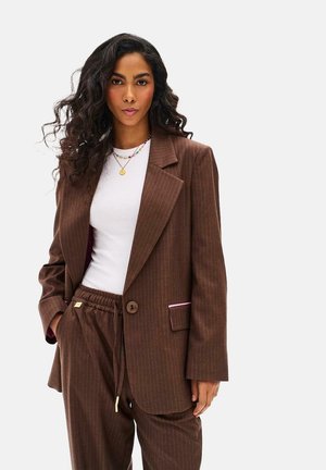 Kobieta o długich kręconych włosach, ubrana w brązową garniturową marynarkę i spodnie w prążki, z białą koszulą i warstwowymi naszyjnikami, stoi z ręką w kieszeni.