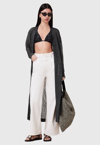 Lång, svart, genomskinlig cardigan över en svart bikiniöverdel, kombinerad med vita högmidjade byxor och svarta sandaler. Accessoarer inkluderar en mönstrad toteväska.