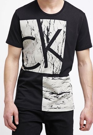 Man draagt een zwart T-shirt met korte mouwen met grote witte blokken met marmerpatroon en zwarte letters "C" en "K" aan de voorkant.