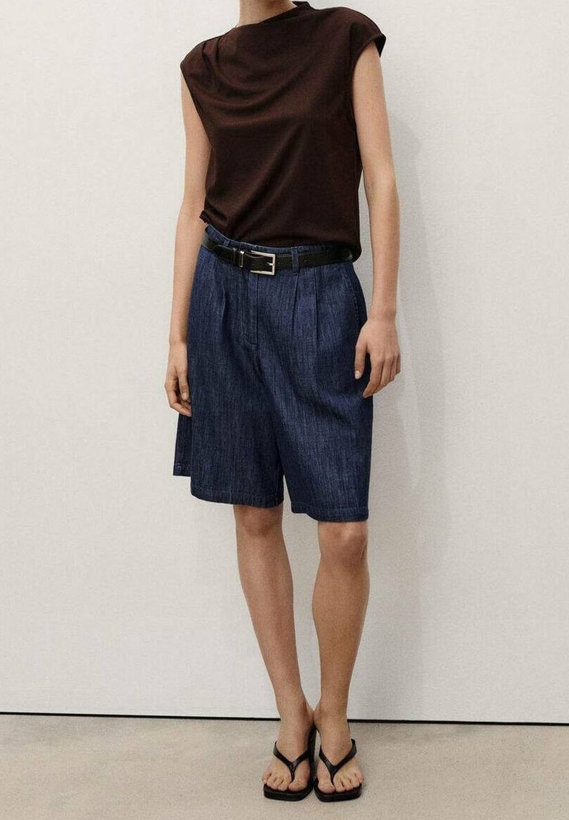 Personne portant un haut marron sans manches, un short bleu foncé taille haute avec ceinture et des sandales noires tongs, debout contre un mur blanc.