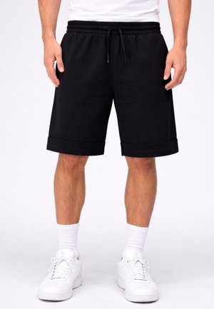 Uomo che indossa pantaloncini neri con coulisse, calzini bianchi e scarpe da ginnastica bianche, in piedi con le mani rilassate lungo i fianchi su sfondo bianco.