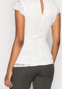 Top bianco in pizzo con maniche a cappuccio, motivo floreale, vita aderente e dettaglio a chiave sulla schiena, abbinato a pantaloni grigio scuro.
