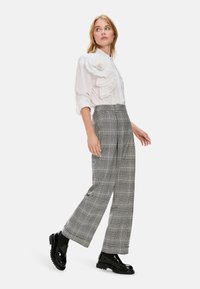 Weißes Blusenshirt mit texturiertem Rüschendetail, kombiniert mit grauen karrierten weiten Hosen. Das Model trägt schwarze Lackloafers.