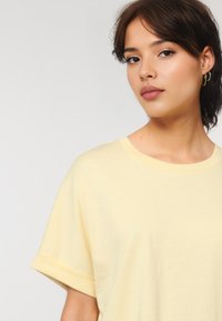 Young woman with short brown hair, wearing a light yellow t-shirt, faces slightly to the side, displaying a neutral expression against a light background.