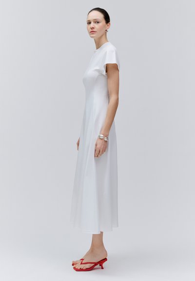 Femme debout portant une robe midi blanche à manches courtes et des sandales rouges à talons, sur un fond gris clair uni.